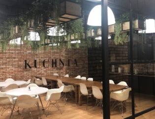 Nowoczesna jadalnia z białymi krzesłami, drewnianymi stołami, ceglanymi ścianami z napisem "KUCHNIA" i wiszącymi roślinami pod sufitem.