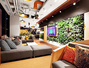 Nowoczesny salon z bujną zieloną ścianą, segmentową sofą, kolorowymi poduszkami i żywą cyfrową ramą artystyczną.