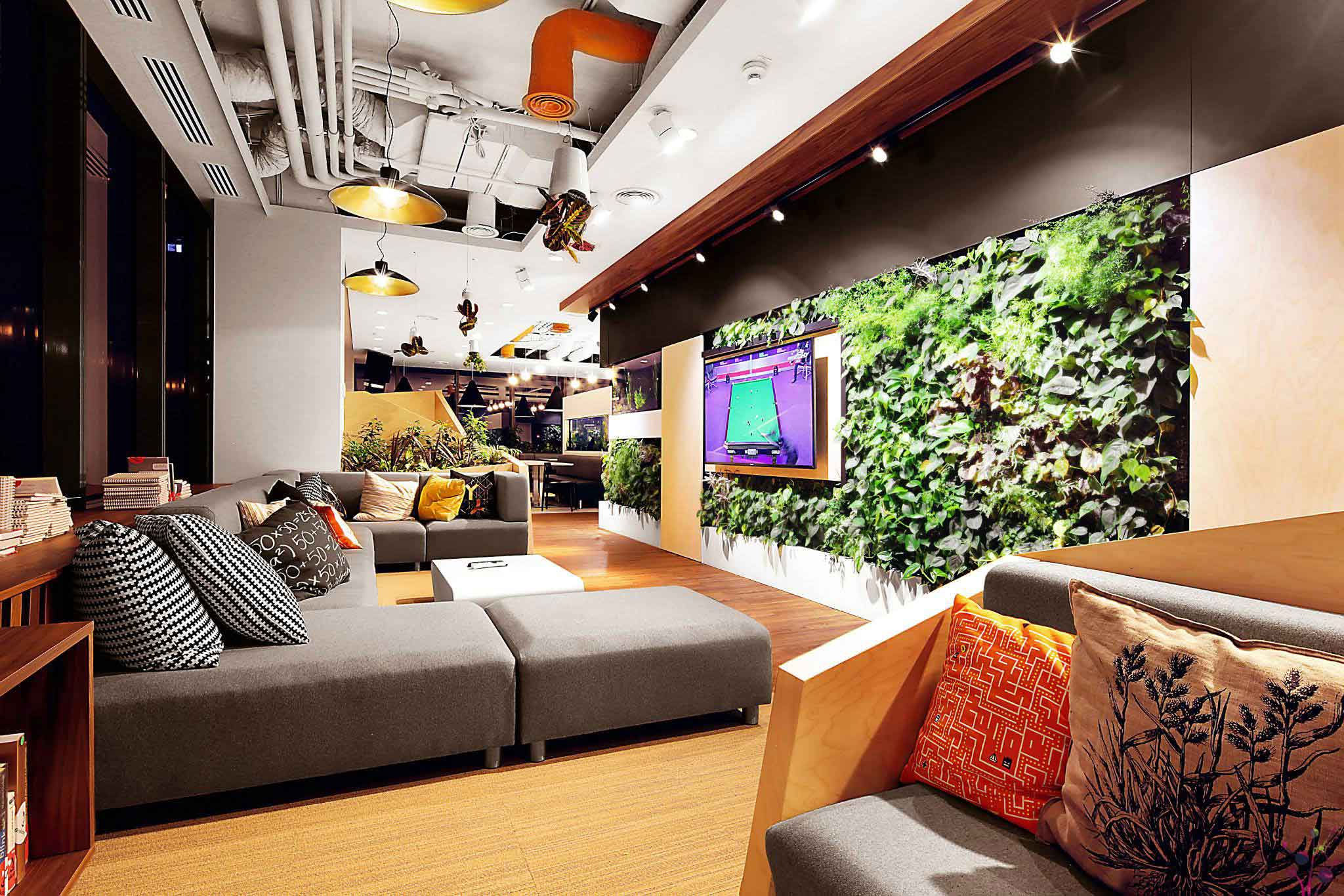 Nowoczesny salon biurowy z szarymi sofami, kolorowymi poduszkami, dużą zieloną żywą ścianą i stylowymi lampami wiszącymi.