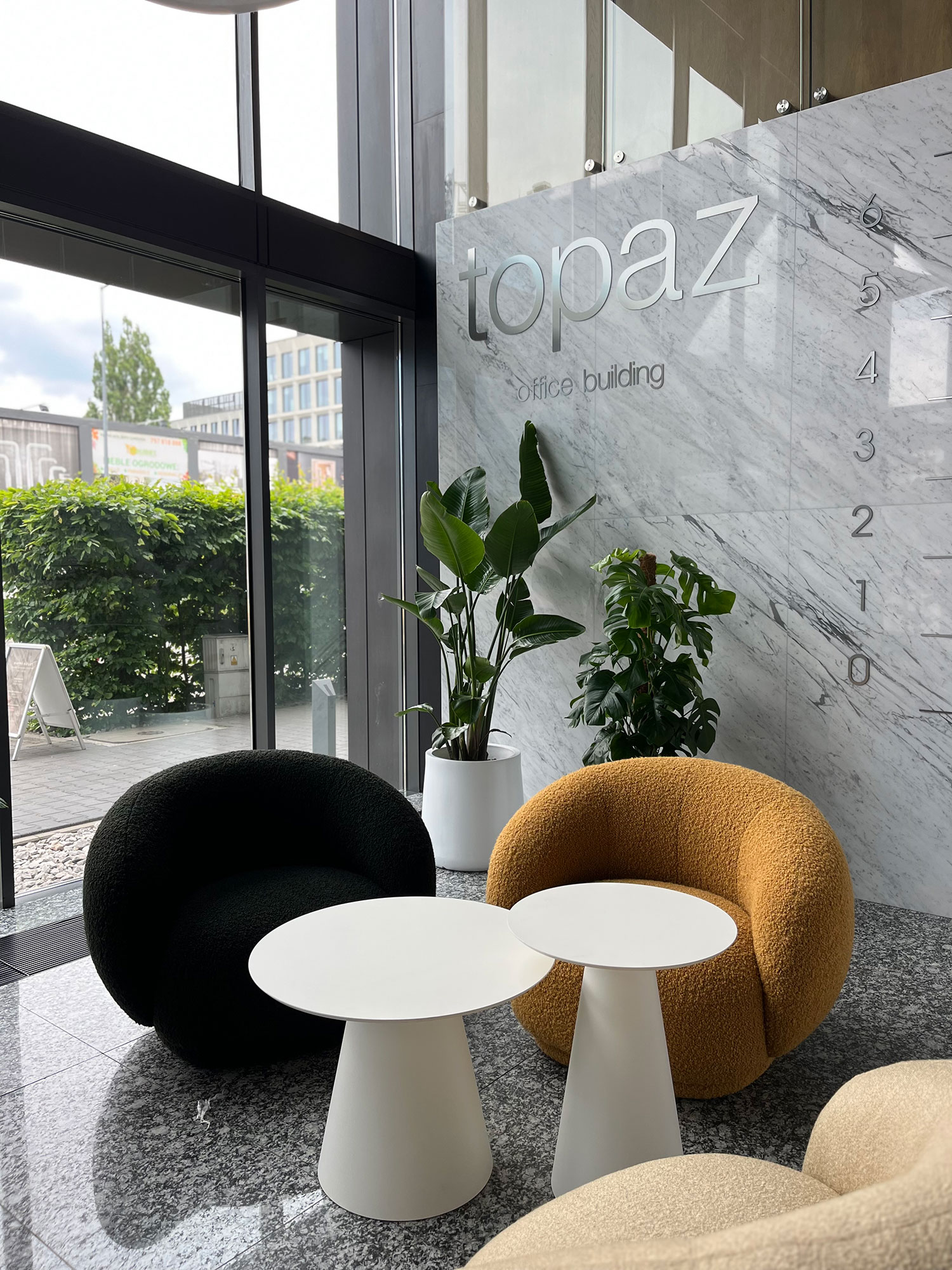 Nowoczesne lobby biurowe „Topaz Office Building” ma marmurową ścianę, dwa czarno-żółte okrągłe krzesła, dwa białe okrągłe stoły i doniczkowe rośliny. Duże okno odsłania bujną zieleń na zewnątrz. To spokojne miejsce pracy, w którym nawet entuzjaści Gry Online znajdują inspirację.