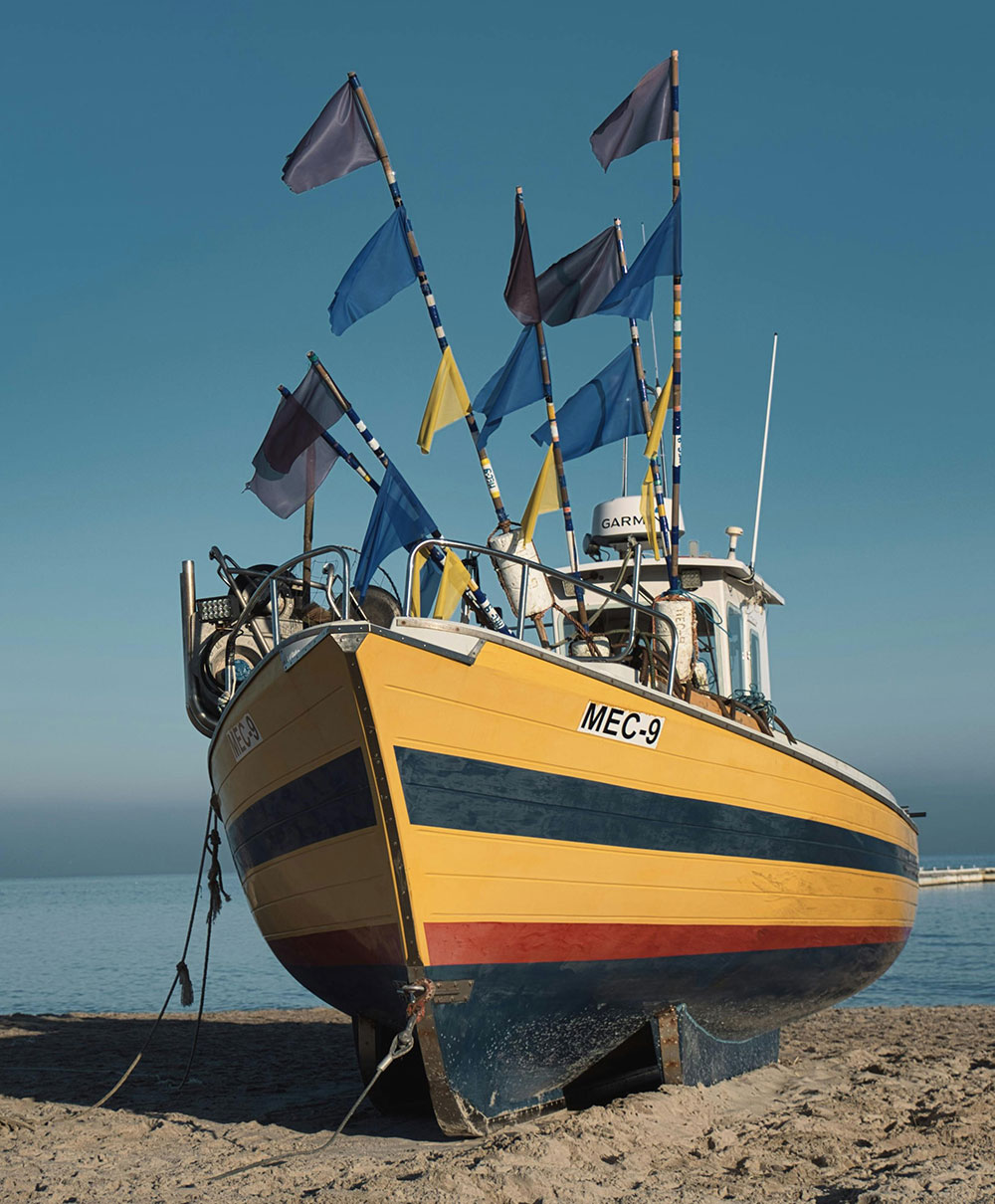 Żółty kuter rybacki o nazwie MEC-9, ozdobiony kolorowymi flagami, spoczywa na piaszczystej plaży niedaleko Gdyni, gdzie równomierna zieleń sprawia, że tętniąca życiem linia brzegowa płynnie łączy się z oceanem i czystym niebem w tle.