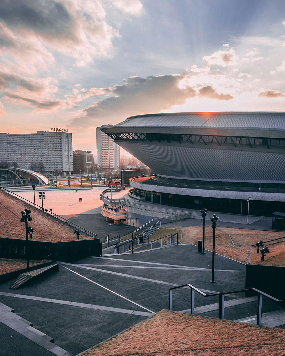 Futurystyczny budynek z zakrzywionym dachem stoi na tle zachodzącego słońca, otoczony miejskim krajobrazem. Szeroki plac i schody prowadzą do wejścia, a ogrody wertykalne dodają odrobinę natury. Zachmurzone niebo pozwala promieniom słonecznym prześwitywać, wzmacniając spokojną atmosferę.