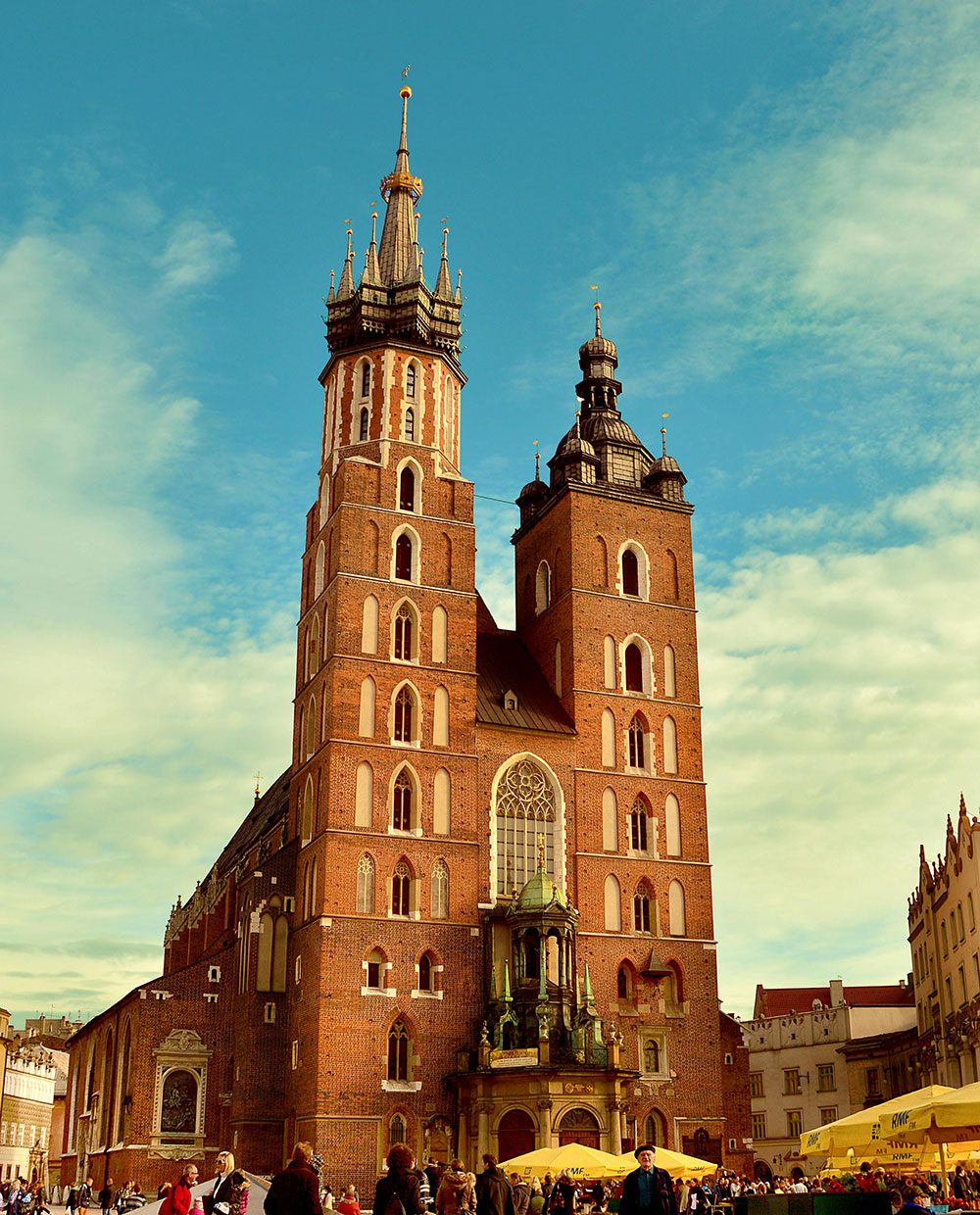 Pod błękitnym niebem majestatycznie wznosi się Bazylika Mariacka z dwiema wieżami, otoczona gwarnymi straganami i tłumami. W pobliżu urok ogrodów wertykalnych dodaje zielonego akcentu do sceny, łącząc naturę z tętniącym życiem miejskim.