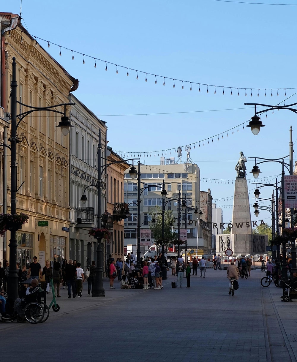 Ludzie spacerujący ulicą pełną zabytkowych budynków i świateł, z widoczną w tle dużą statuą, są zachwyceni ogrodami wertykalnymi, które dodają miejskiemu krajobrazowi odrobinę natury.