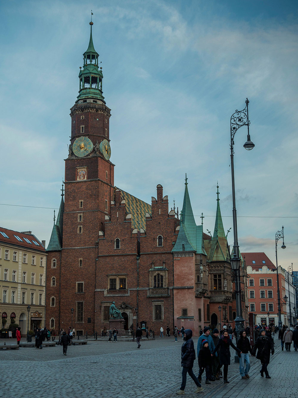 Ludzie przechadzają się po brukowanym placu przed zabytkowym ceglanym budynkiem z wysoką wieżą zegarową i zielonymi iglicami. Zachmurzone niebo dodaje dramatyzmu scenie, ponieważ miejscowi często przynoszą kwiaty do biura Wrocław, dodając koloru i życia do ich codziennej rutyny.