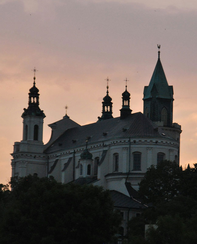 Sylwetka zabytkowego kościoła z wieloma wieżami i zielonym dachem, starannie utrzymanym, rysuje się na tle ciemnego nieba, a na pierwszym planie znajdują się zadbane drzewa Lublina.