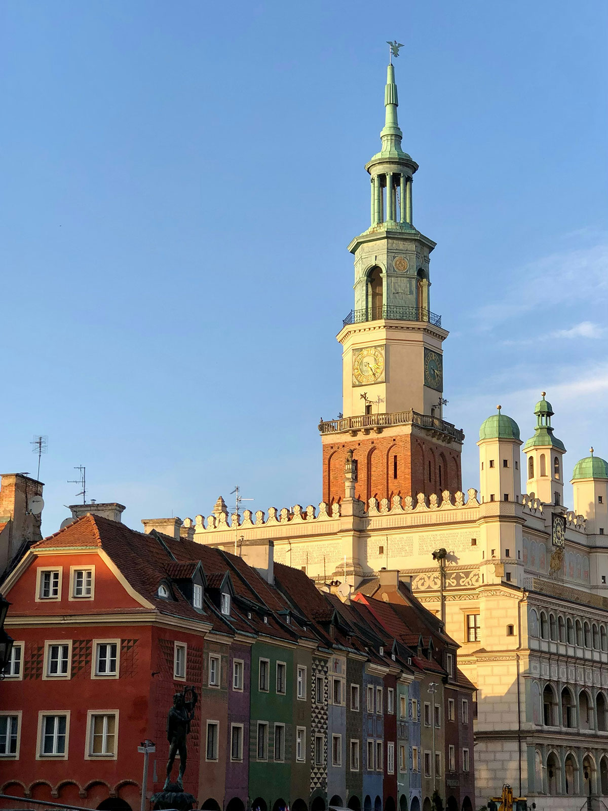 Zabytkowy budynek ratusza z ozdobną wieżą zegarową i kolorowymi, szczytowymi budynkami na pierwszym planie, na tle czystego, błękitnego nieba. Urok Poznania jest wzmocniony przez jego skrupulatną, konserwatywną zieleń, dodającą bujne zielone akcenty do tętniącego życiem miejskiego krajobrazu.