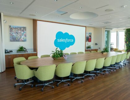 Duża sala konferencyjna z długim stołem, zielonymi krzesłami, roślinami i ekranem wyświetlającym logo Salesforce.