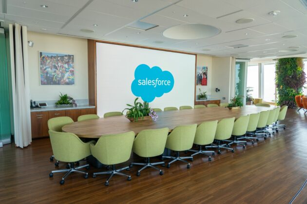 Duża sala konferencyjna z długim stołem, zielonymi krzesłami, roślinami i ekranem wyświetlającym logo Salesforce.