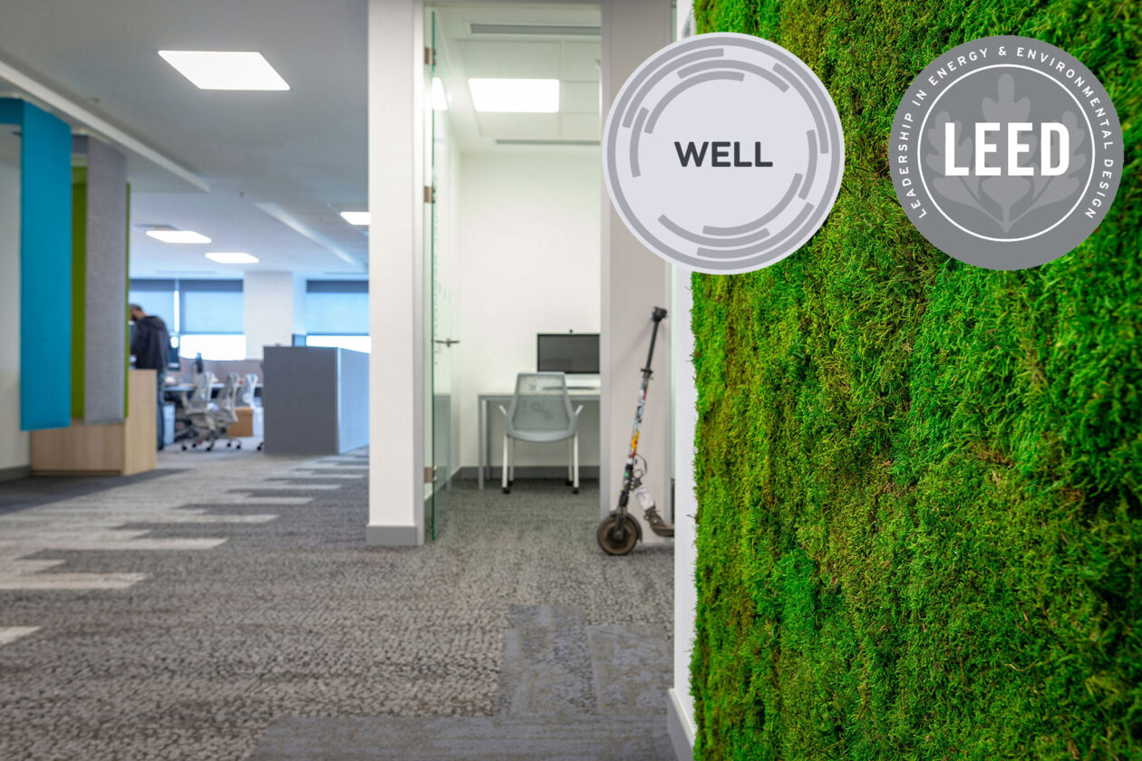 Nowoczesne wnętrze biurowe ze ścianą pokrytą zielonym mchem, logo certyfikatów WELL i LEED, widocznymi stanowiskami pracy i skuterem przy przeszklonym biurze.