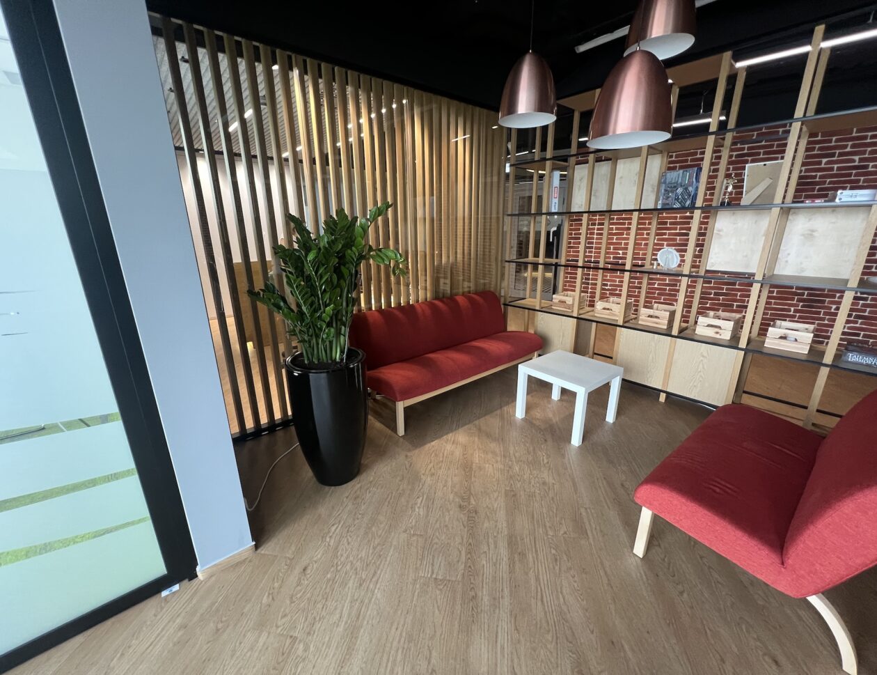 Nowoczesny salon biurowy z czerwonymi sofami, białym stolikiem kawowym, dużą rośliną doniczkową, drewnianą podłogą, regałem i drewnianą ścianką działową.
