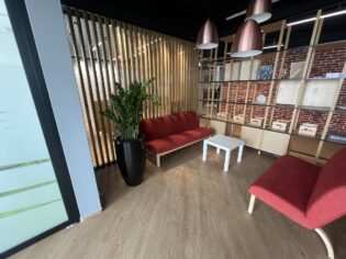 Nowoczesny salon biurowy z czerwonymi sofami, białym stolikiem kawowym, dużą rośliną doniczkową, drewnianą podłogą, regałem i drewnianą ścianką działową.