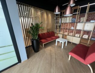 Nowoczesny salon biurowy z czerwonymi sofami, białym stolikiem kawowym, dużą rośliną doniczkową, drewnianą podłogą, regałem i drewnianą ścianką działową.