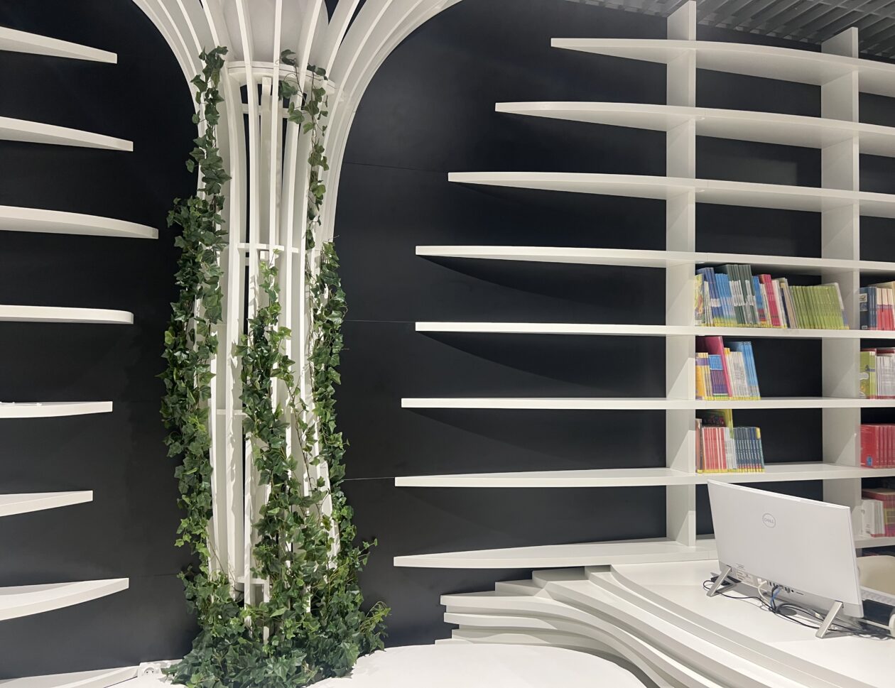 Nowoczesne wnętrze biblioteki z białymi zakrzywionymi półkami na książki, komputerem na biurku i kolumną owiniętą zielonym bluszczem; niektóre książki ułożone według kolorów na prawych półkach.
