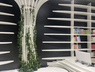 Nowoczesne wnętrze biblioteki z białymi zakrzywionymi półkami na książki, komputerem na biurku i kolumną owiniętą zielonym bluszczem; niektóre książki ułożone według kolorów na prawych półkach.