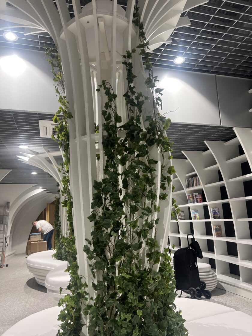 Zakrzywione białe półki na książki i kolumny architektoniczne z zielonymi winoroślami w nowoczesnej przestrzeni bibliotecznej; osoba pracuje w tle.