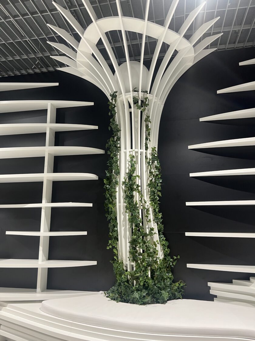 Biała nowoczesna struktura architektoniczna z zakrzywionymi pionowymi listwami, półkami po obu stronach i zielonymi pnączami wspinającymi się po środku, na czarnym tle.