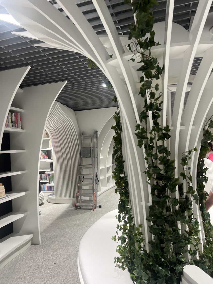 Nowoczesne wnętrze biblioteki z zakrzywionymi białymi półkami na książki, metalową drabiną i pnącymi się zielonymi winoroślami na centralnej białej strukturze.