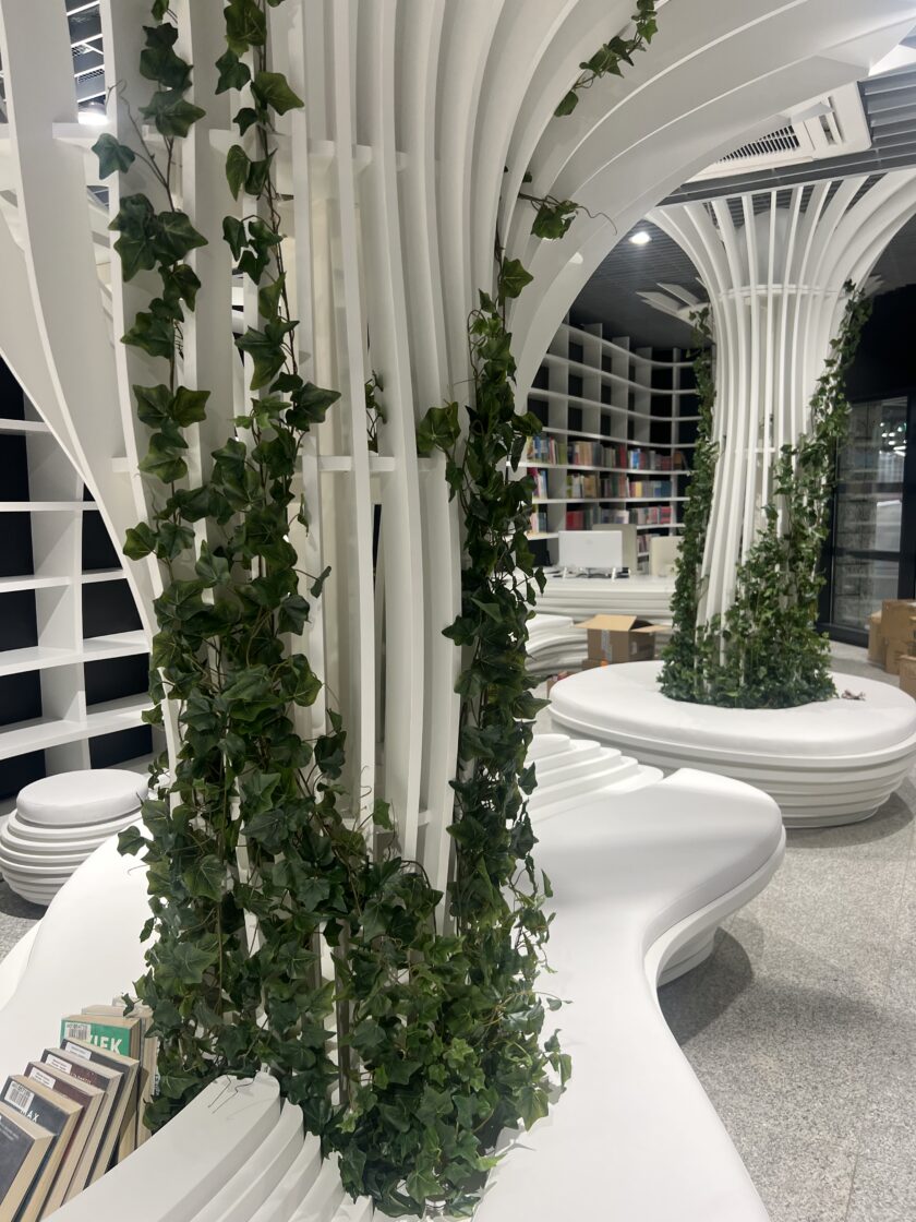 Nowoczesne wnętrze biblioteki z zakrzywionymi białymi półkami i siedzeniami, z kolumnami owiniętymi zieloną winoroślą i książkami ułożonymi na półkach w tle.