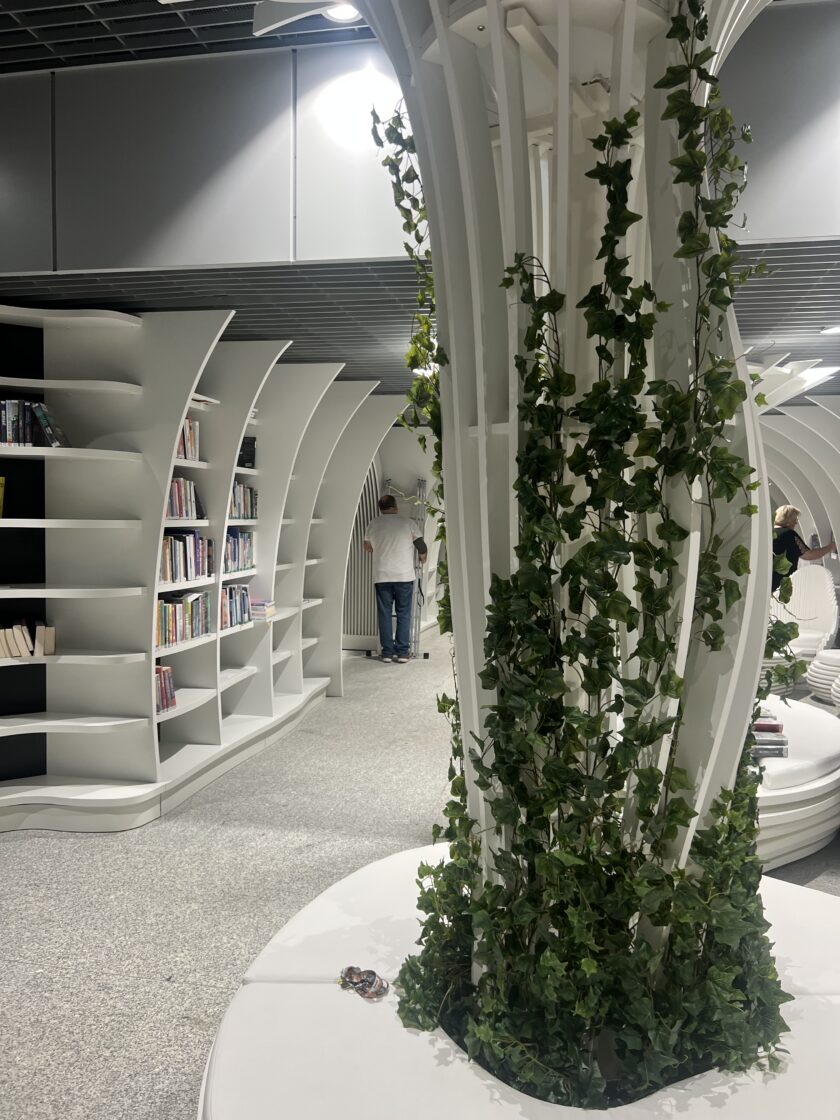Nowoczesne wnętrze biblioteki z zakrzywionymi białymi półkami na książki, pionowym wspornikiem owiniętym zielonym bluszczem i osobą przeglądającą książki w tle.