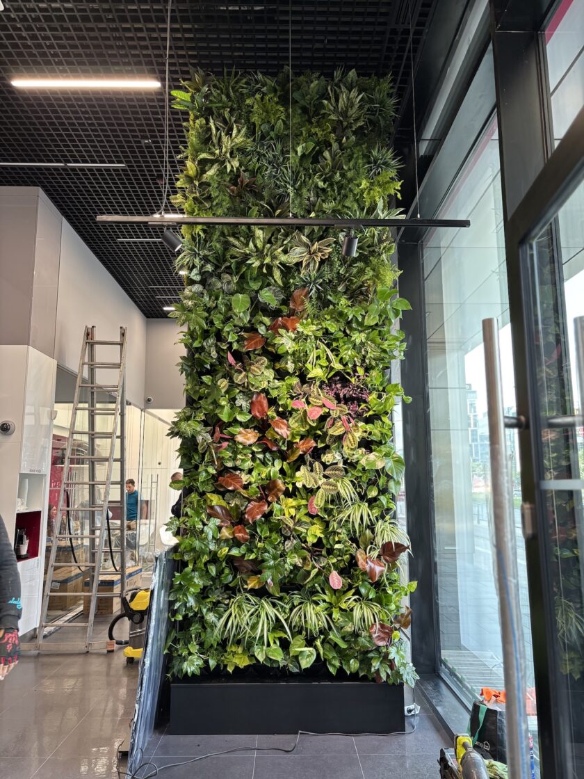 Wysoki pionowy ogród wewnętrzny z różnymi zielonymi i czerwonymi roślinami został zainstalowany w nowoczesnym holu budynku, obok dużych okien i metalowej drabiny.