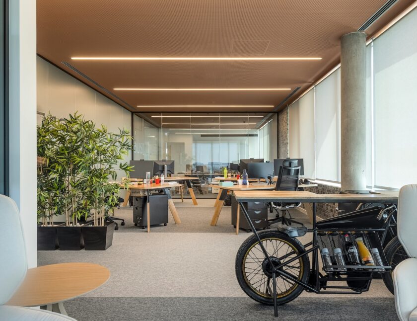 Nowoczesne biuro na otwartym planie z biurkami, krzesłami biurowymi, roślinami doniczkowymi, dużymi oknami z żaluzjami i czarnym rowerem z butelkami wina na pierwszym planie.