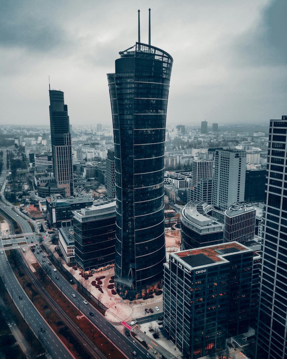 Wysoki, ciemny, nowoczesny wieżowiec stoi wśród innych budynków w miejskim krajobrazie pod zachmurzonym niebem, a kwiaty do biura Warszawa dodają elegancji do tętniącej życiem atmosfery.