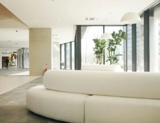 Przestronne nowoczesne lobby z dużymi oknami, białymi zakrzywionymi sofami, wysokimi roślinami i lampami wiszącymi. W tle widoczne są dwie osoby przy ścianie.