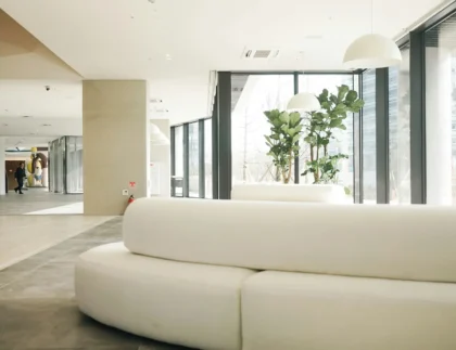 Przestronne nowoczesne lobby z dużymi oknami, białymi zakrzywionymi sofami, wysokimi roślinami i lampami wiszącymi. W tle widoczne są dwie osoby przy ścianie.
