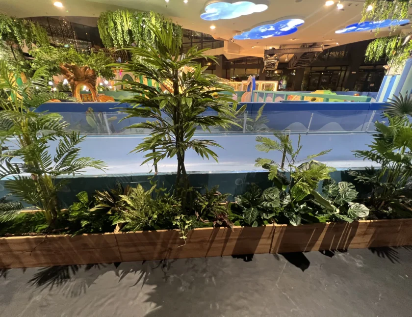 Drewniana skrzynka wypełniona różnymi zielonymi roślinami stoi przed niebieską barierką, a w tle znajduje się jasno oświetlony wewnętrzny plac zabaw.