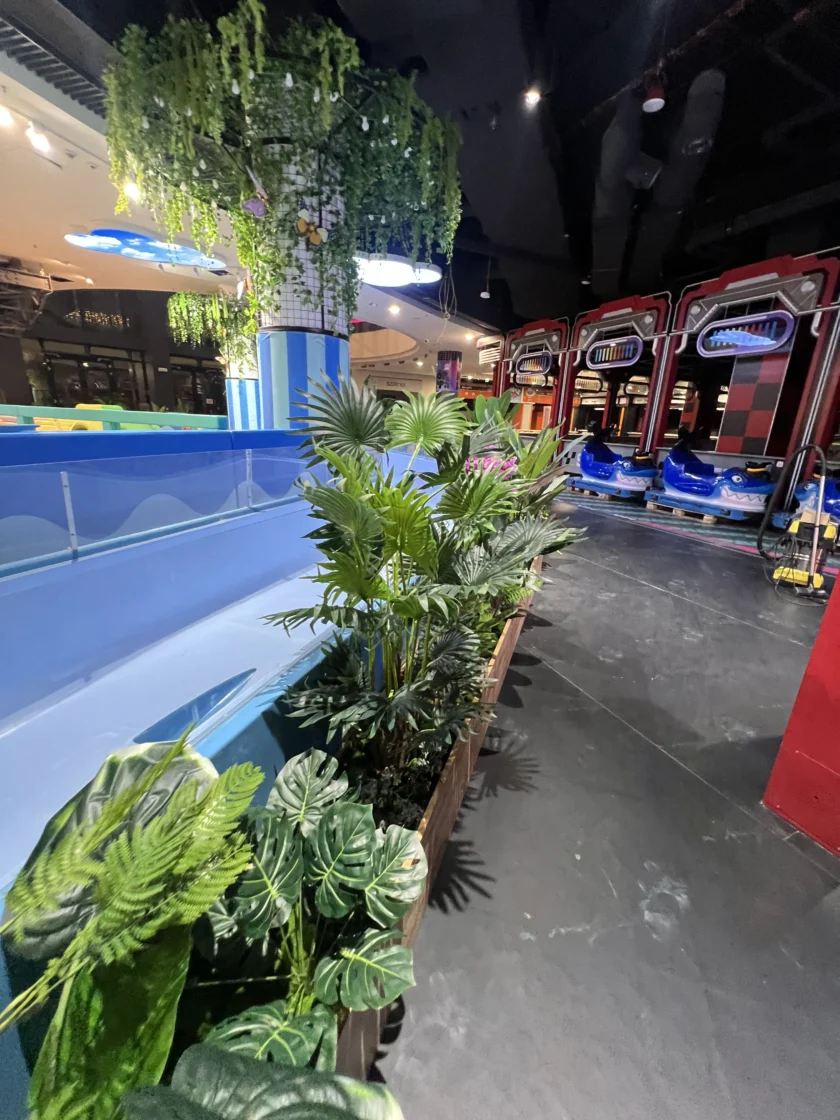 Kryta strefa rozrywki z samochodzikami w tle, zielonymi roślinami doniczkowymi na pierwszym planie i sztuczną zielenią zwisającą z sufitu.
