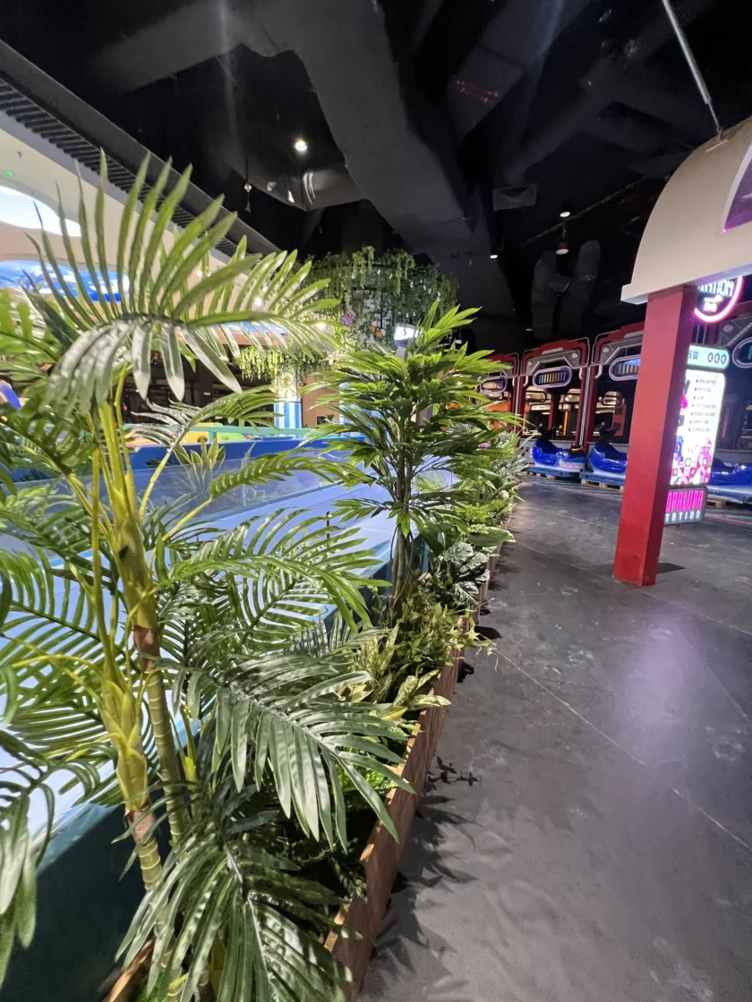 Kryta strefa rozrywki ze sztucznymi roślinami na pierwszym planie, sekcją wyłożoną niebieską wykładziną i widocznymi atrakcjami w postaci samochodzików zderzakowych w tle.