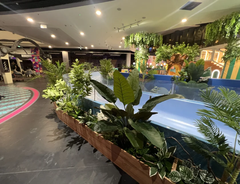 Obszar wewnętrzny z zielonymi roślinami doniczkowymi w drewnianej donicy, niebieską balustradą i elementami dekoracyjnymi, w tym posągiem żyrafy i wiszącymi roślinami.