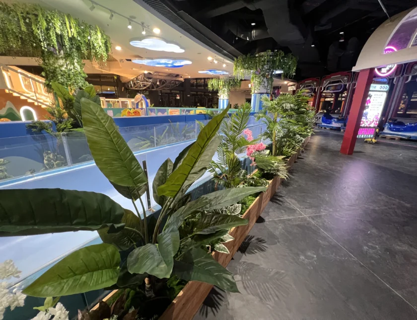 Kryta strefa rozrywki z grami zręcznościowymi w tle, sztucznymi roślinami wyściełającymi niebieską przegrodę i dekoracyjnym oświetleniem nad głową.