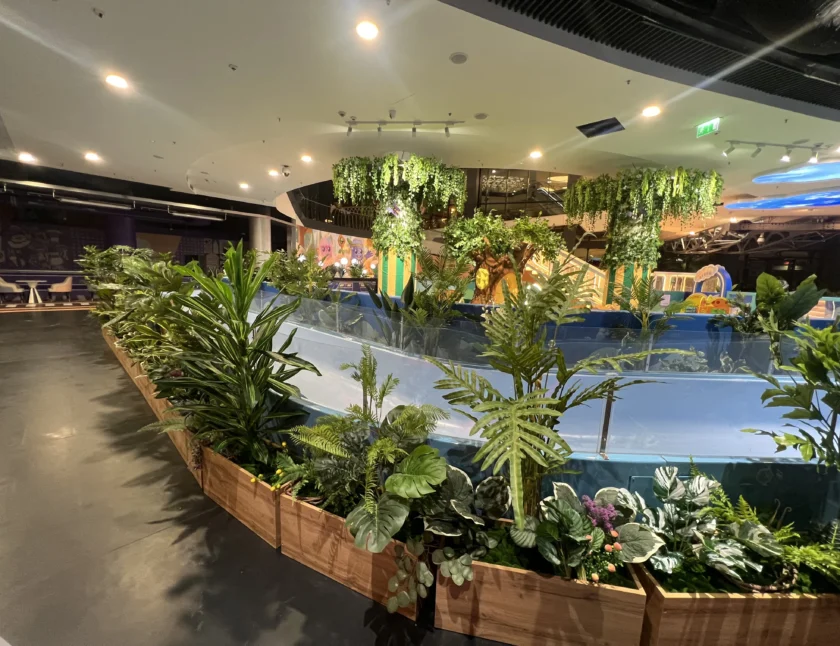 Wewnętrzny ogród z różnymi zielonymi roślinami w drewnianych donicach otacza niebieski chodnik; sufit ma wiszącą zieleń i światła, a kolorowy plac zabaw jest widoczny w tle.