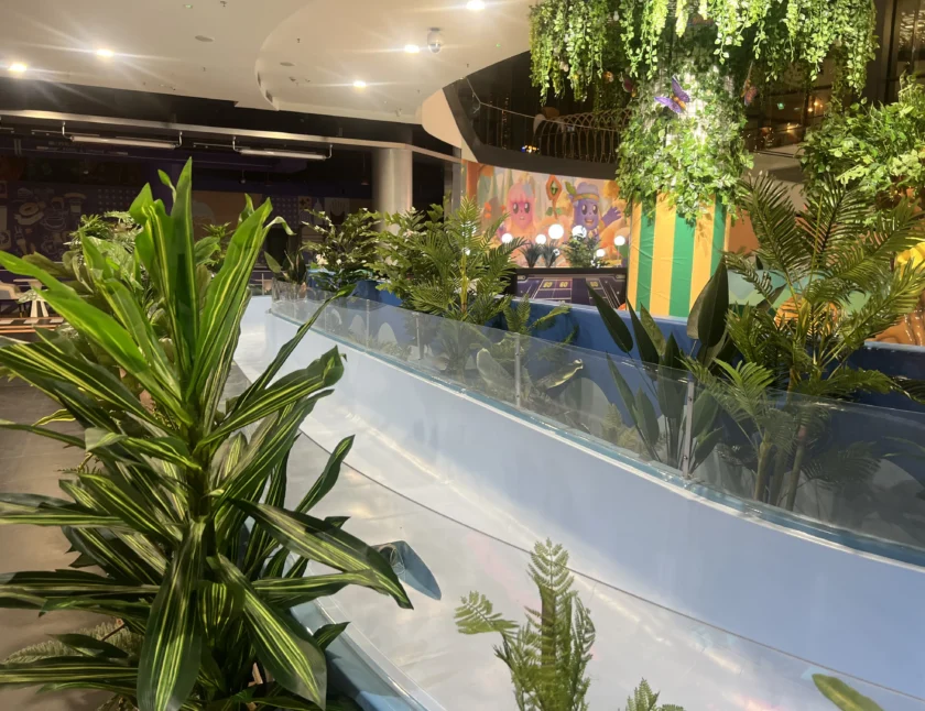 Przestrzeń wewnętrzna ze sztucznymi roślinami wzdłuż niebieskiej zakrzywionej bariery, jasnym oświetleniem i kolorowymi dekoracjami ściennymi w tle.