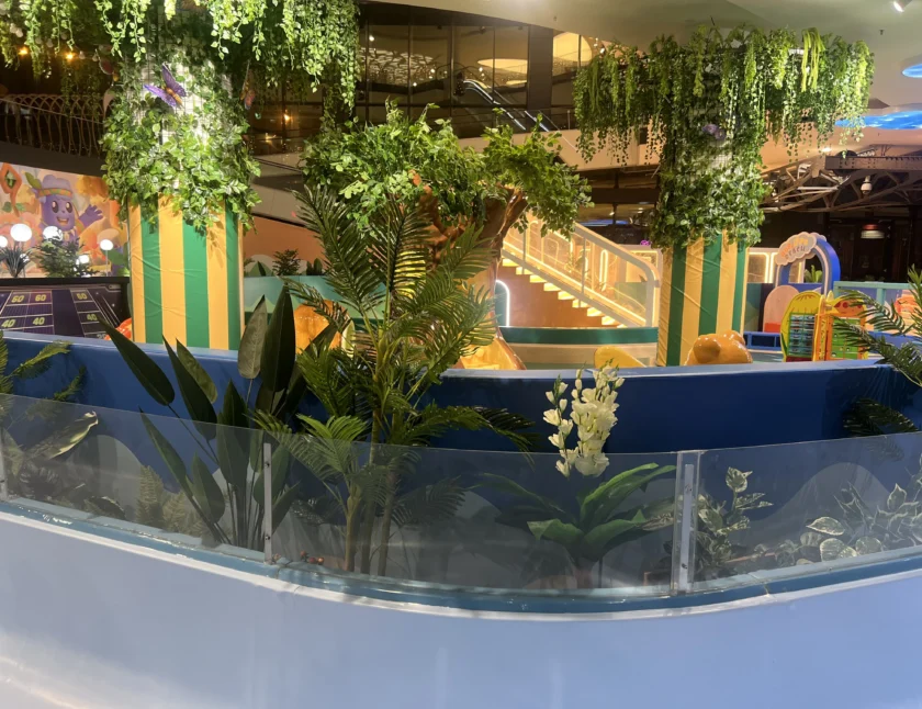 Wewnętrzny plac zabaw z bujnymi zielonymi roślinami, schodami z żółtymi światłami i kolorowymi dekoracjami w tle.