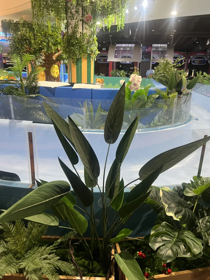 Kryta strefa rozrywki ze sztucznymi roślinami na pierwszym planie, niebieską balustradą i kolorowymi dekoracjami, w tym drzewem i figurkami zwierząt w tle.