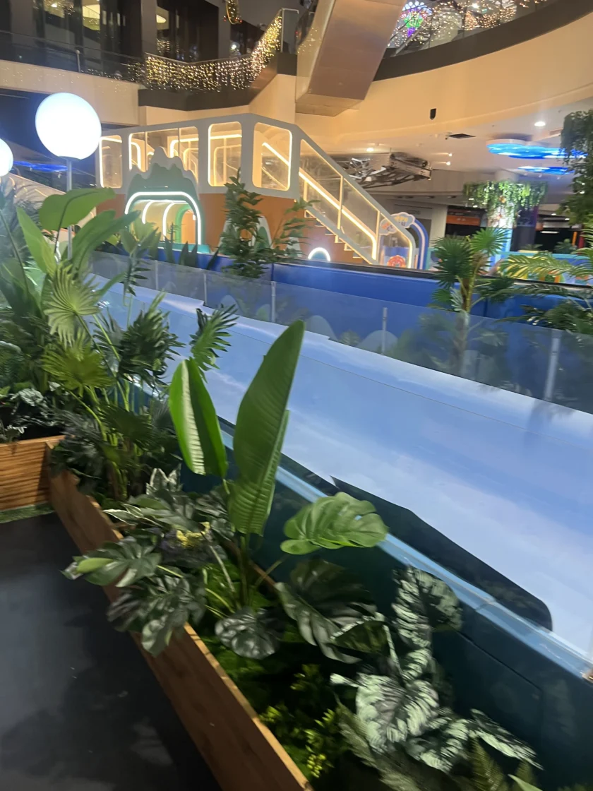 Scena w pomieszczeniu z zielonymi roślinami doniczkowymi na pierwszym planie i niebieską, przeszkloną zjeżdżalnią lub rampą z podświetlanymi schodami i dekoracjami w tle.