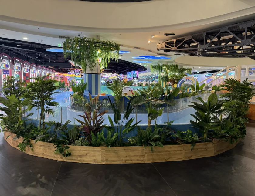 Wewnętrzny ogród z różnymi zielonymi roślinami w drewnianej donicy, położony wewnątrz nowoczesnego centrum handlowego lub kompleksu rozrywkowego z kolorowymi światłami w tle.