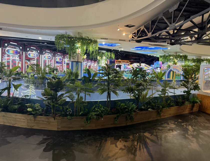 Obszar wewnętrzny z różnymi zielonymi roślinami w drewnianej donicy; w tle widoczne są kolorowe światła, znaki i gry zręcznościowe.