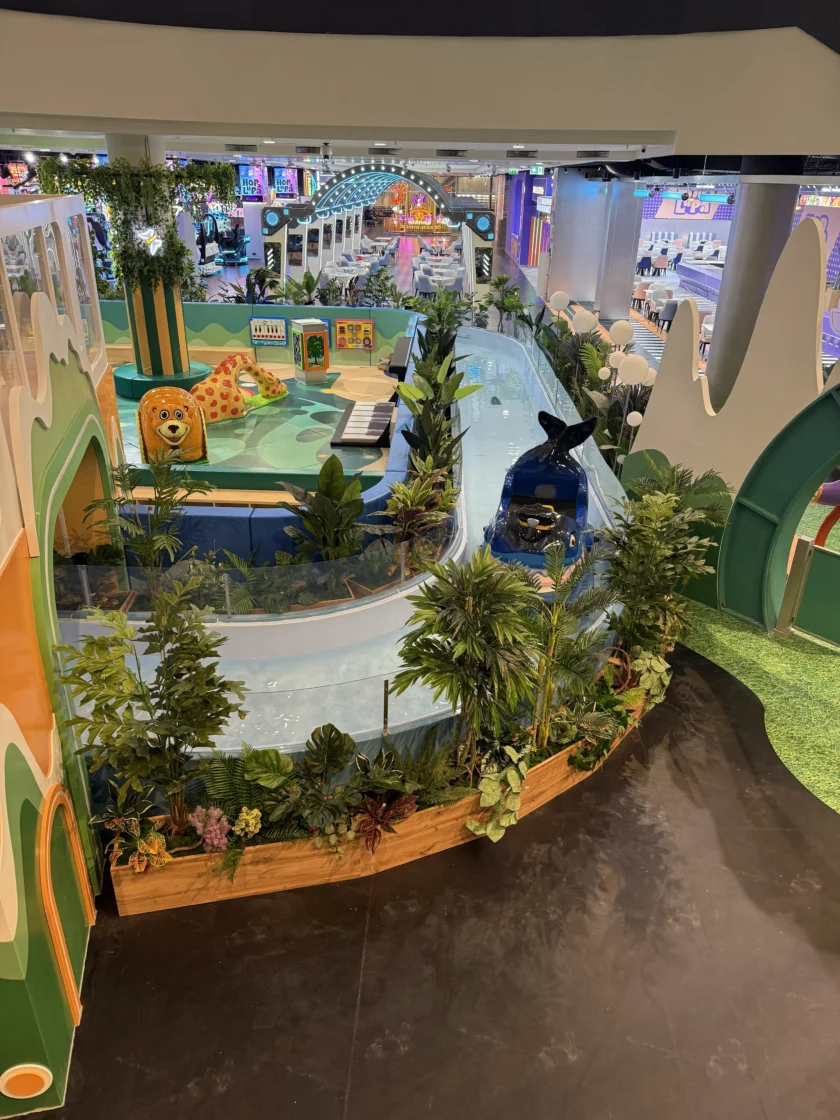 Kryty plac zabaw dla dzieci z płytkim, krętym kanałem wodnym, sztucznymi roślinami, dekoracjami o tematyce zwierzęcej i kolorowymi konstrukcjami do zabawy.