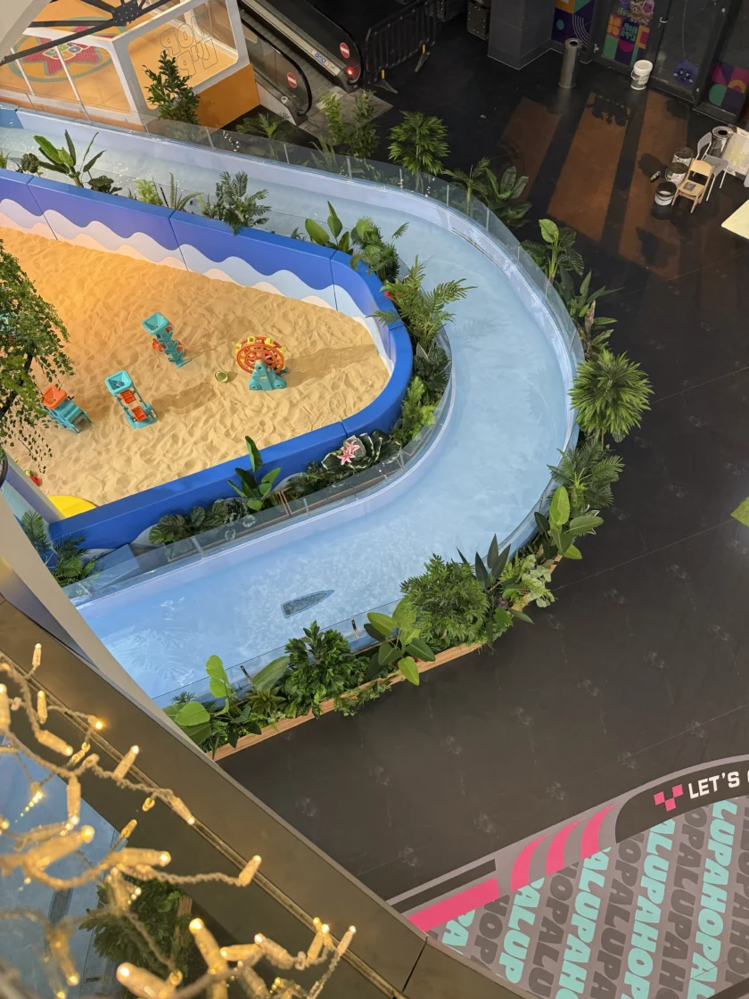 Wewnętrzny plac zabaw obejmuje kręty, płytki kanał wodny otaczający piaskownicę z zabawkowymi przedmiotami plażowymi, otoczony roślinami i dekoracjami.