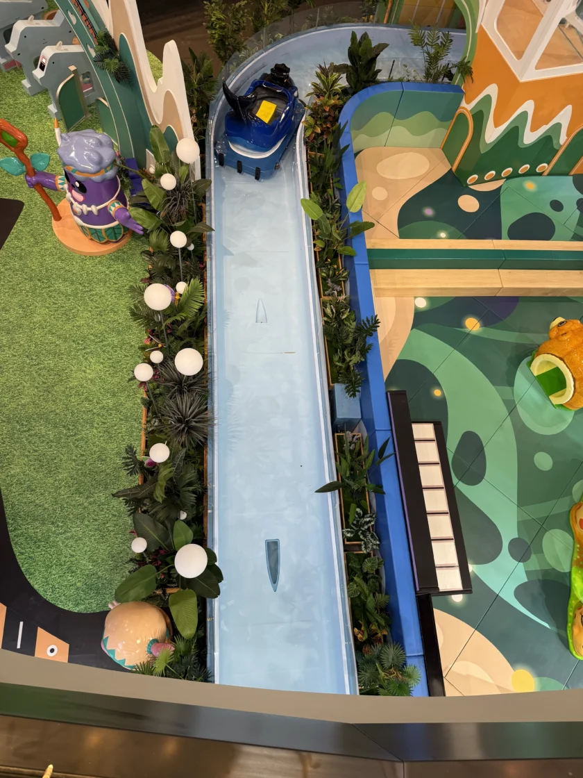 Widok z lotu ptaka na niebieską łódź z bali w płytkim kanale wodnym otoczonym kolorowymi konstrukcjami zabawowymi i sztuczną zielenią.