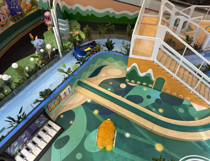 Kolorowy wewnętrzny plac zabaw ze zjeżdżalnią, konstrukcjami zwierząt, dużym fortepianem, roślinami i placem zabaw z krętą ścieżką i dekoracyjnymi wzorami.