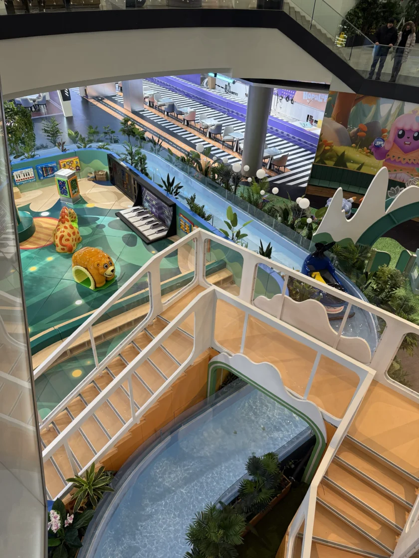 Kryty plac zabaw z zabawkami w kształcie zwierząt, suchą zjeżdżalnią i miejscem do siedzenia w nowoczesnym centrum handlowym. Widoczne są różne poziomy i zieleń.
