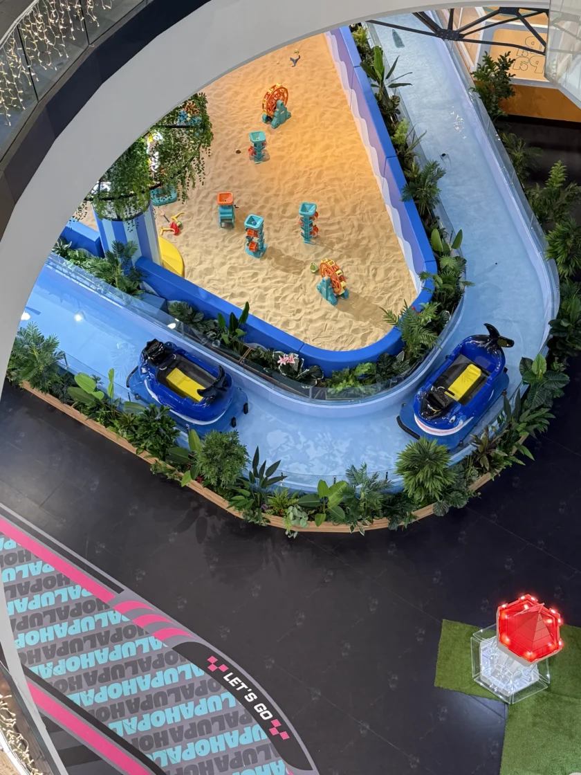 Widok z góry na wewnętrzny plac zabaw z piaszczystą sekcją z zabawkowymi zwierzętami i otaczającym ją torem dla samochodów elektrycznych z dwoma niebieskimi samochodami.