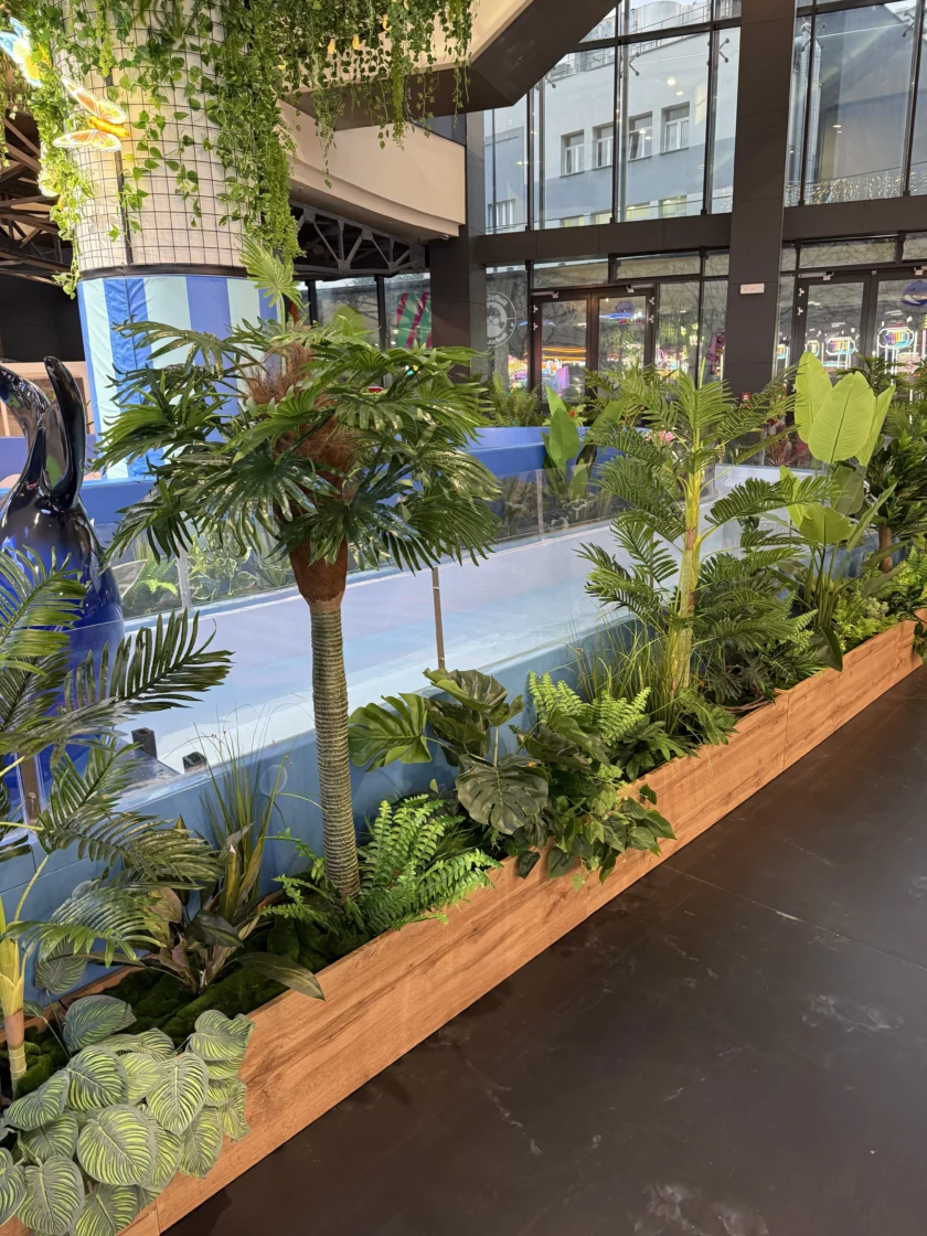 Drewniana donica wypełniona różnymi zielonymi sztucznymi roślinami stoi przed przezroczystą barierą w pobliżu krytego lodowiska.