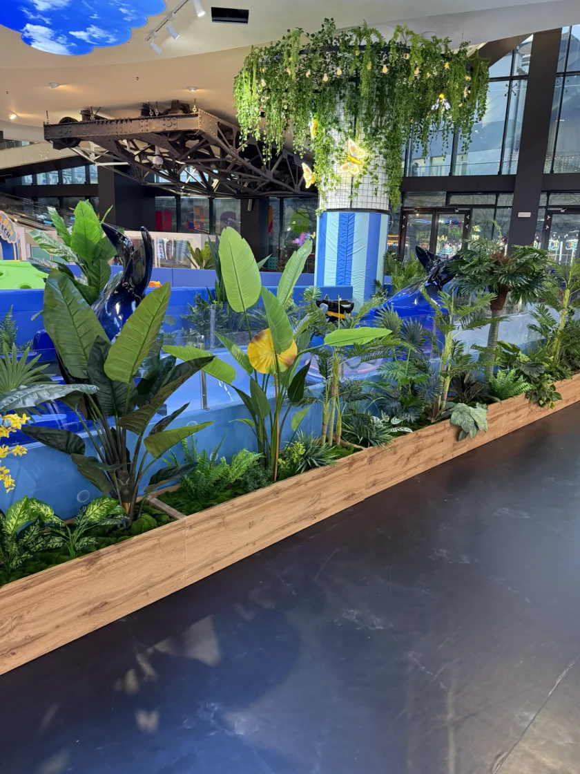 Scena w pomieszczeniu przedstawiająca drewnianą donicę z różnymi tropikalnymi roślinami przed niebieskim placem zabaw, z zielenią zwisającą z filaru w tle.