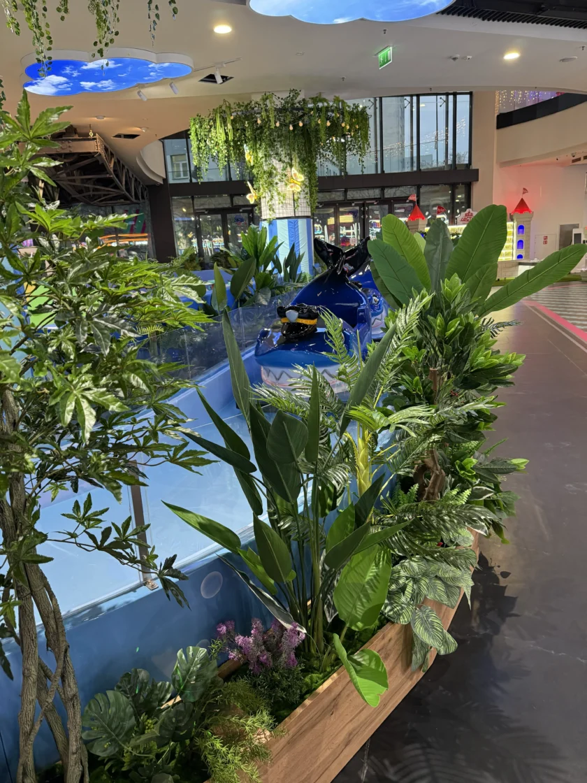 Wewnętrzna scena z bujnymi zielonymi roślinami na pierwszym planie, niebieskim wyświetlaczem skutera wodnego za szkłem i nowoczesną przestrzenią handlową w tle.
